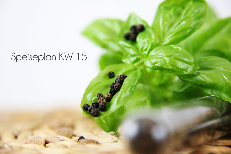 Ein frisches Basilikumblatt mit schwarzen Pfefferkörnern ist im Vordergrund sichtbar. Im Hintergrund steht der Text „Speiseplan KW 15“. Der Untergrund ist eine geflochtene Oberfläche.