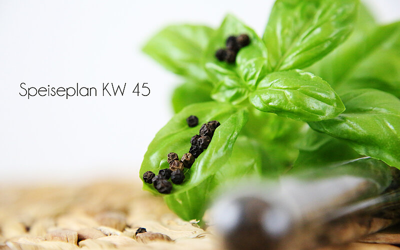 Ein grünes Basilikumblatt mit einigen schwarzen Pfefferkörnern ist im Vordergrund zu sehen. Im Hintergrund steht in ansprechender Schrift "Speiseplan KW 45". Der untere Bereich zeigt eine gewebte Unterlage.