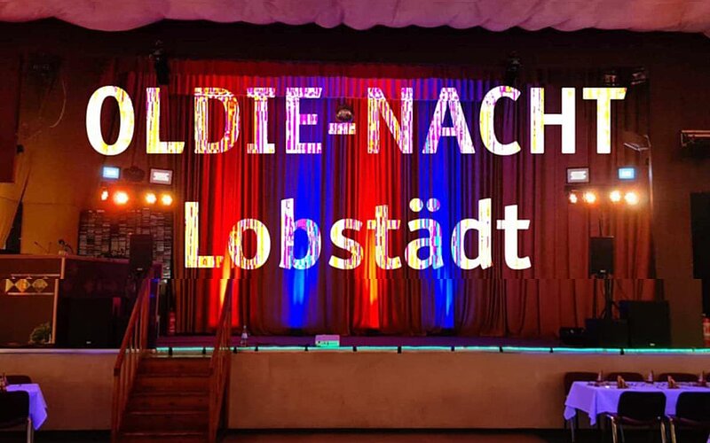 Ein festlich dekorierter Raum mit rotem und blauem Vorhang im Hintergrund. In großen, bunten Buchstaben steht „OLDIE-NACHT Lobstädt“ im Vordergrund. Es gibt eine Bühne mit Beleuchtung und einem Bühnenaufbau, das auf eine Veranstaltung hinweist.