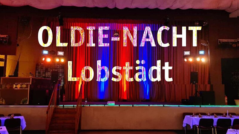 Ein festlich dekorierter Raum mit rotem und blauem Vorhang im Hintergrund. In großen, bunten Buchstaben steht „OLDIE-NACHT Lobstädt“ im Vordergrund. Es gibt eine Bühne mit Beleuchtung und einem Bühnenaufbau, das auf eine Veranstaltung hinweist.