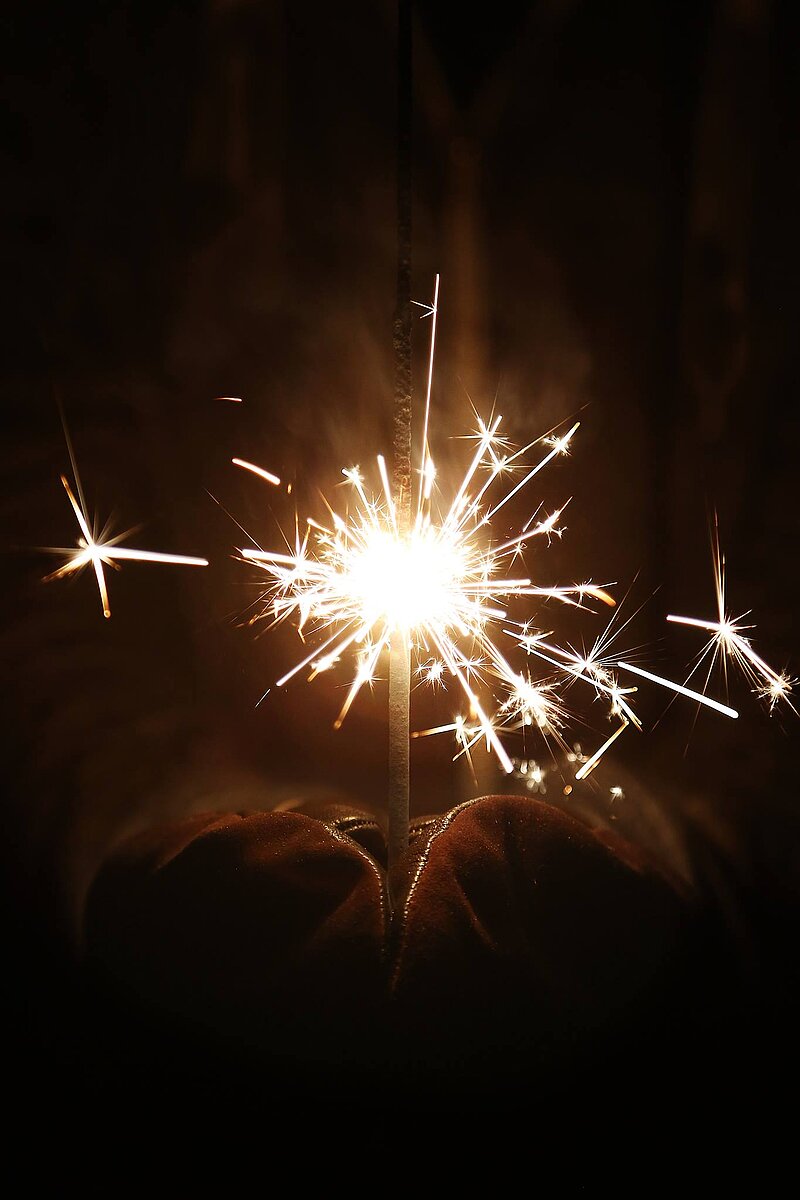 Eine Hand hält einen brennenden Feuerwerkskörper, der helle Funken und Licht erzeugt. Der Hintergrund ist dunkel, wodurch die Strahlen des Feuerwerks besonders leuchten.