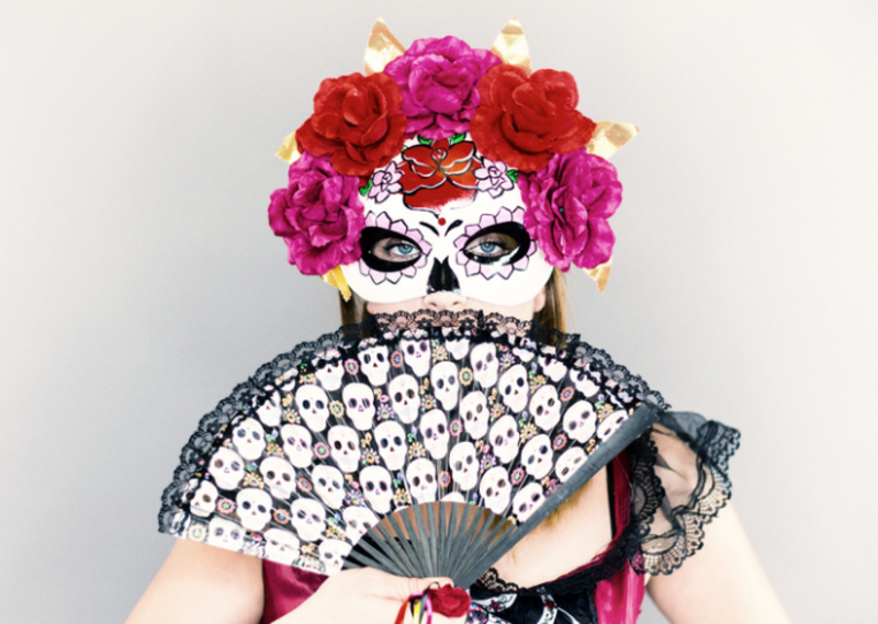 Eine Person trägt eine verzierte Totenschädelmaske mit bunten Blumen und hält einen Fächer mit Schädelmotiven. Der Hintergrund ist neutral, was die auffälligen Farben und Details der Maske und des Fächers betont.