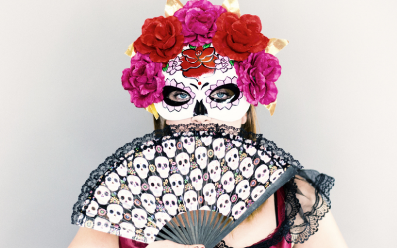 Eine Person trägt eine verzierte Totenschädelmaske mit bunten Blumen und hält einen Fächer mit Schädelmotiven. Der Hintergrund ist neutral, was die auffälligen Farben und Details der Maske und des Fächers betont.