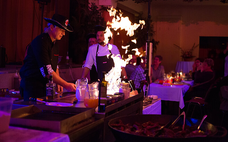 Zwei Köche stehen an einer Grillstation, während eine Flamme hochschießt. Einer der Köche trägt einen Hut und arbeitet mit Zutaten auf der Arbeitsfläche. Im Hintergrund sind Tische mit Gästen zu sehen, die das Geschehen aufmerksam beobachten. Die Beleuchtung ist gedämpft.
