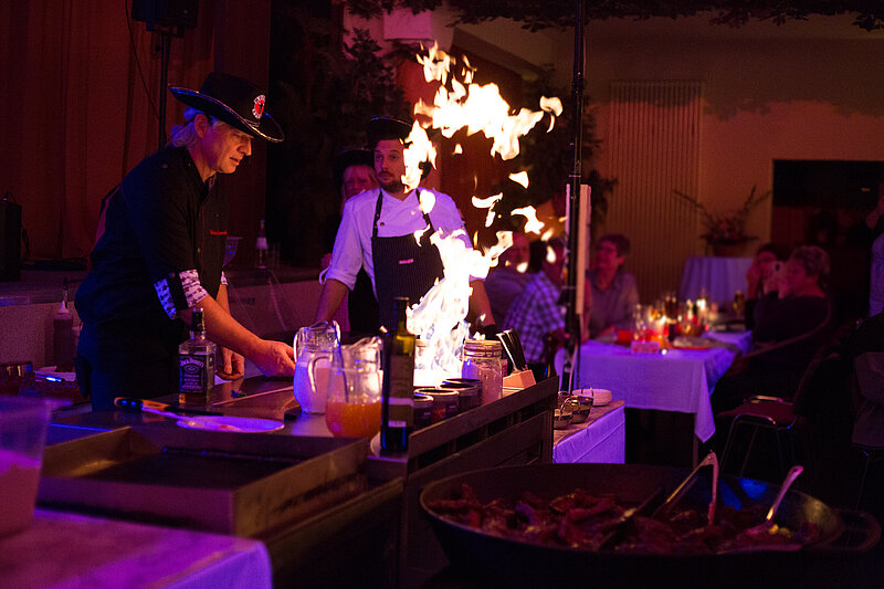Zwei Köche stehen an einer Grillstation, während eine Flamme hochschießt. Einer der Köche trägt einen Hut und arbeitet mit Zutaten auf der Arbeitsfläche. Im Hintergrund sind Tische mit Gästen zu sehen, die das Geschehen aufmerksam beobachten. Die Beleuchtung ist gedämpft.